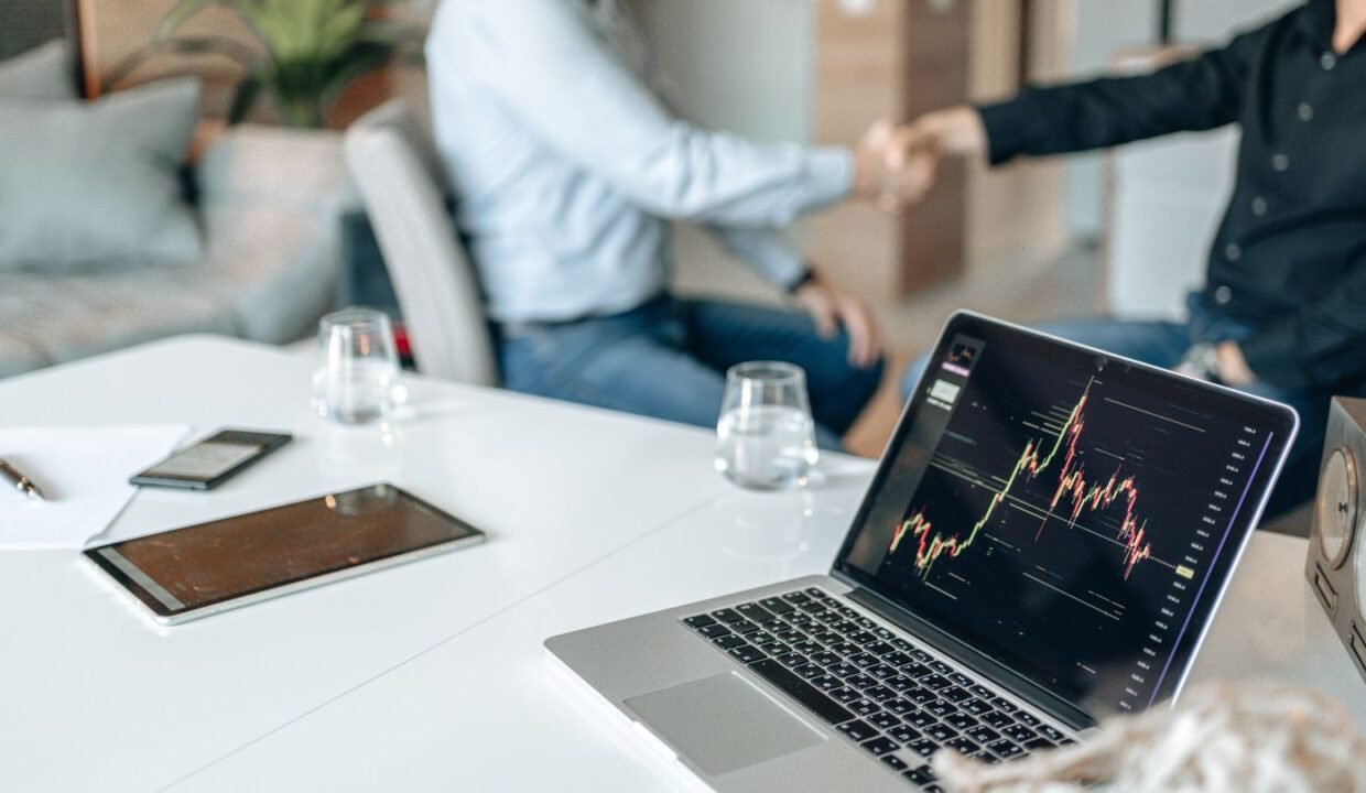 Two professionals shaking hands in an office with data analysis on a laptop screen