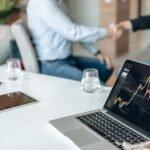 Two professionals shaking hands in an office with data analysis on a laptop screen