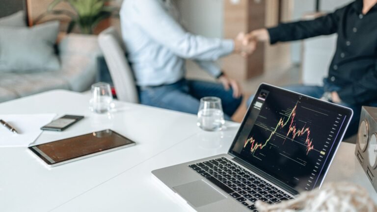 Two professionals shaking hands in an office with data analysis on a laptop screen
